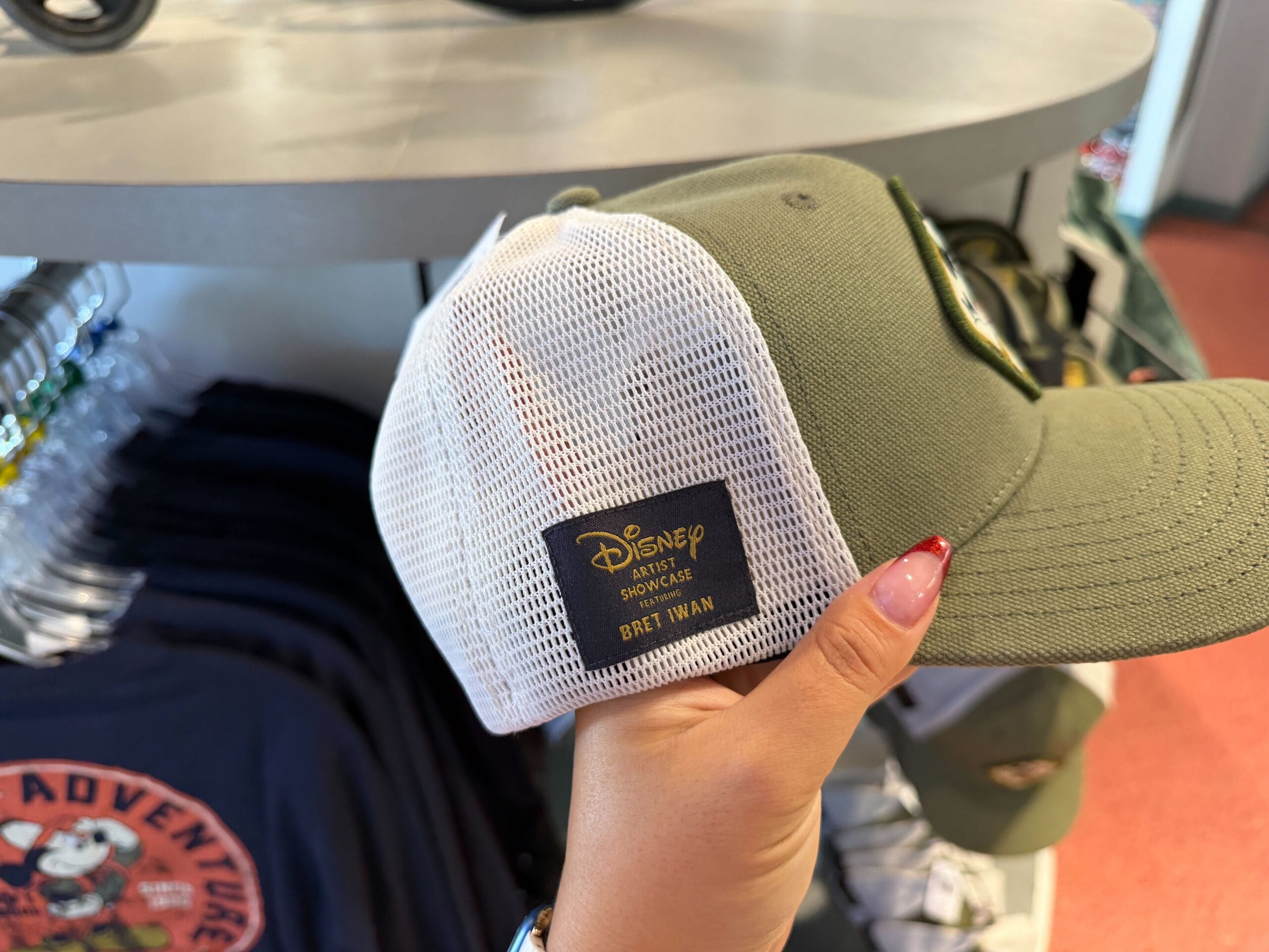 A hand holds an olive green and white Disney hat from the Expedition Mickey Mouse collection, labeled "Bret Iwan," at a Disney park.