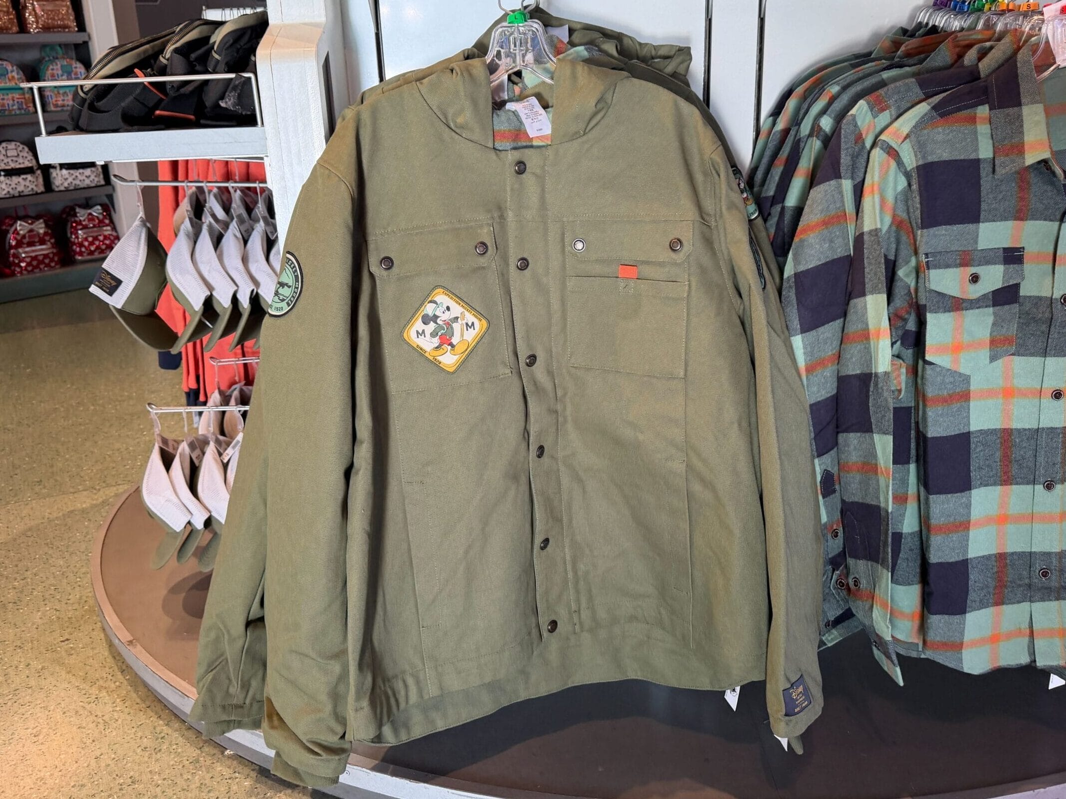 Two long-sleeve Expedition Mickey Mouse shirts hang on a rack in a Disney Park shop, surrounded by magical park merchandise.