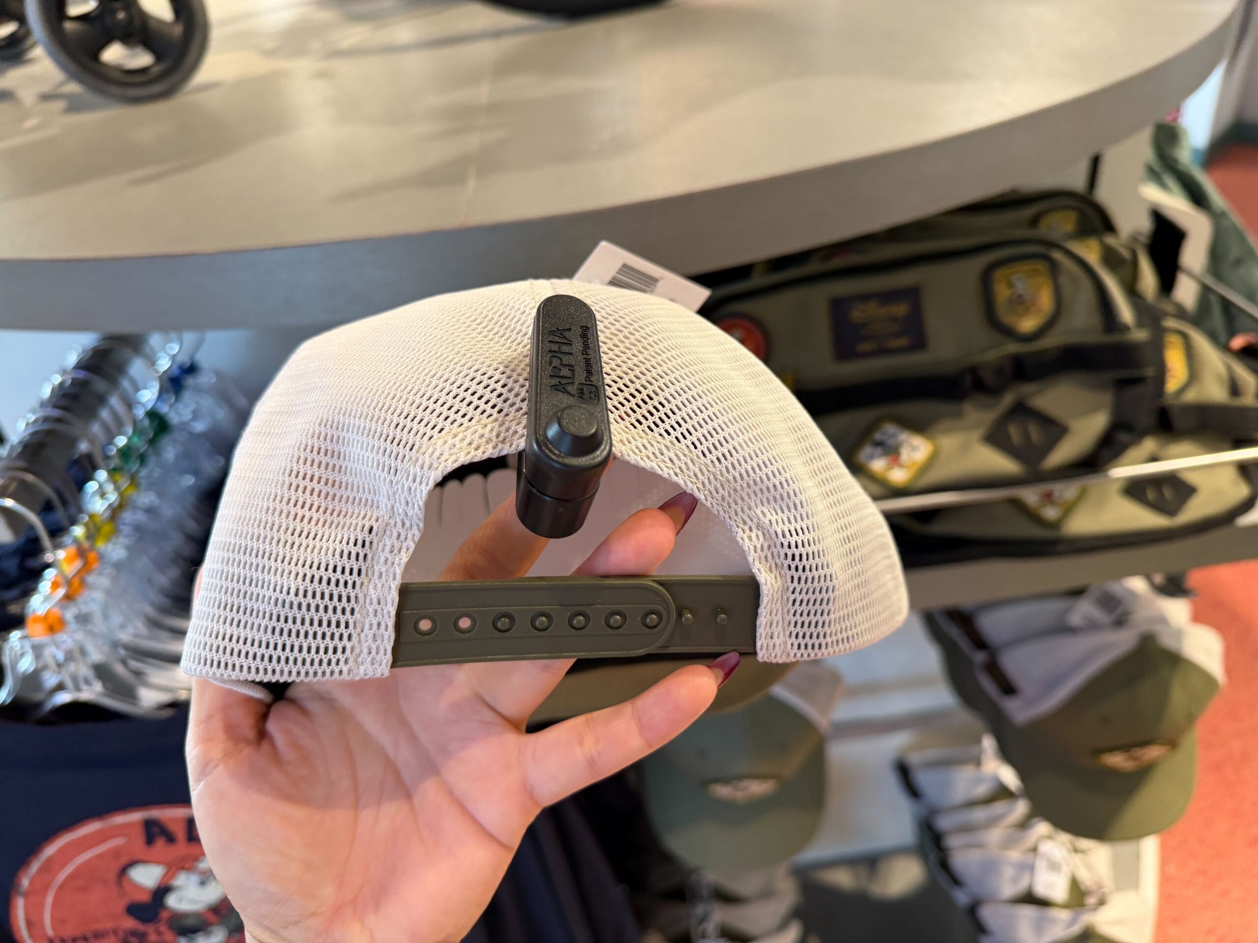 A hand holds a white mesh-back hat with a security tag; shelves behind display Expedition Mickey Mouse hats and bags, Disney Parks style.
