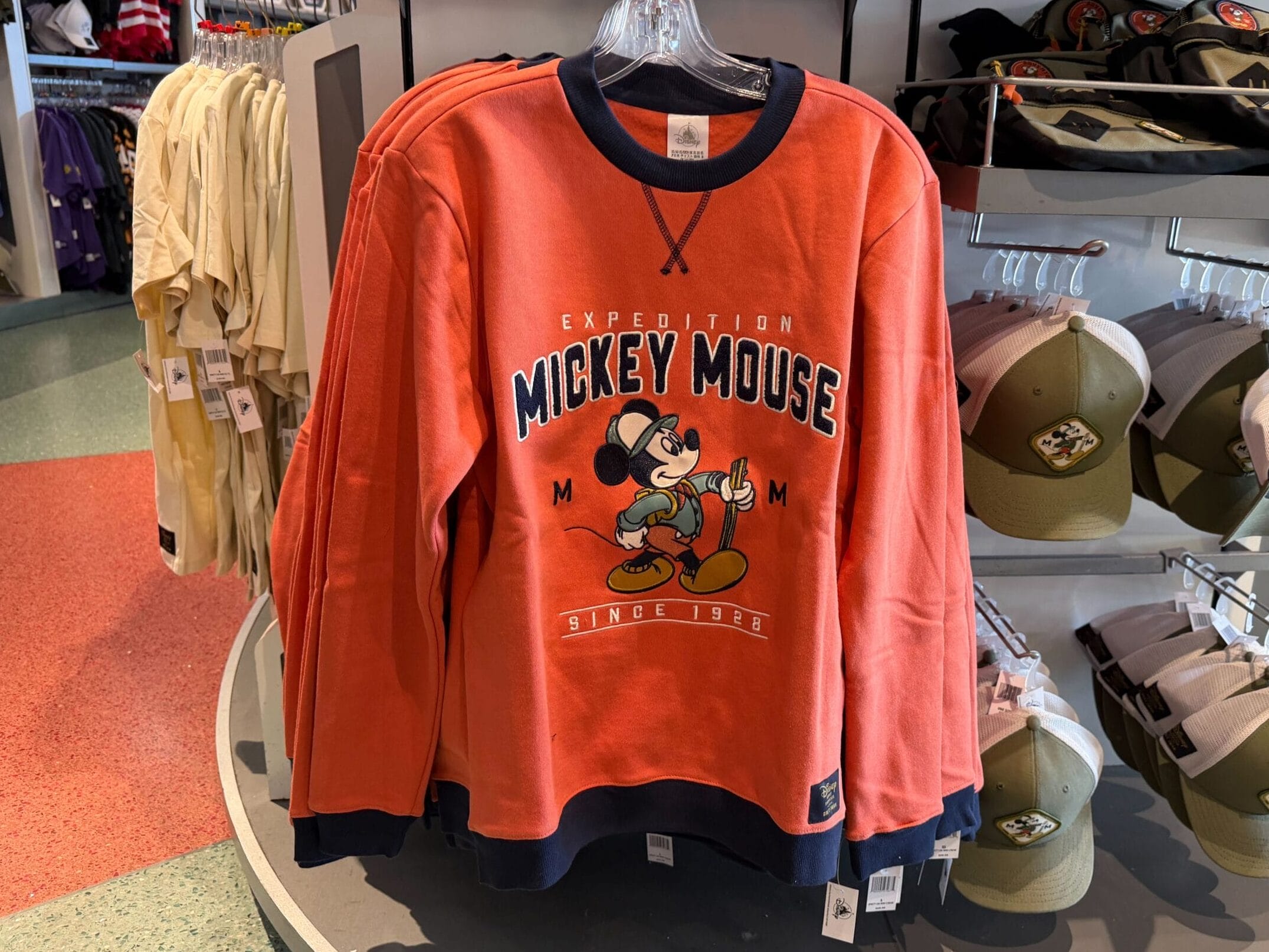 Orange “Mickey Mouse” sweatshirt from the Expedition Mickey Mouse Collection, shown in a Disney Park shop display with signage.