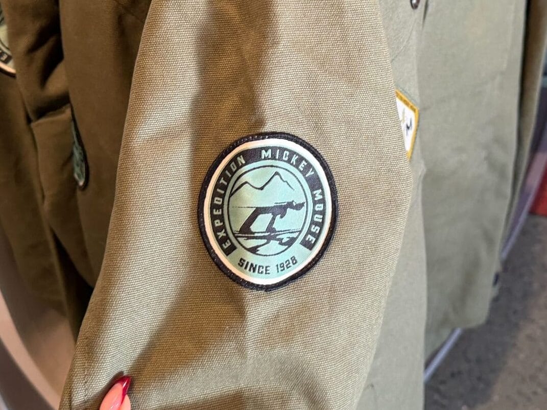 Guest holds an olive green Expedition Mickey Mouse jacket, styled with park flair, among similar jackets in a Disney store.