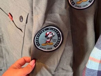 A hand displays a green Expedition Collection shirt featuring Mickey Mouse, with two matching shirts hanging nearby, perfect Disney park style.