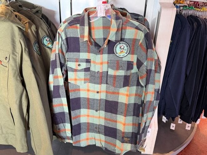 A green and gray plaid flannel shirt from the Mickey Mouse Collection with a cartoon bird patch on display at a Disney park store.
