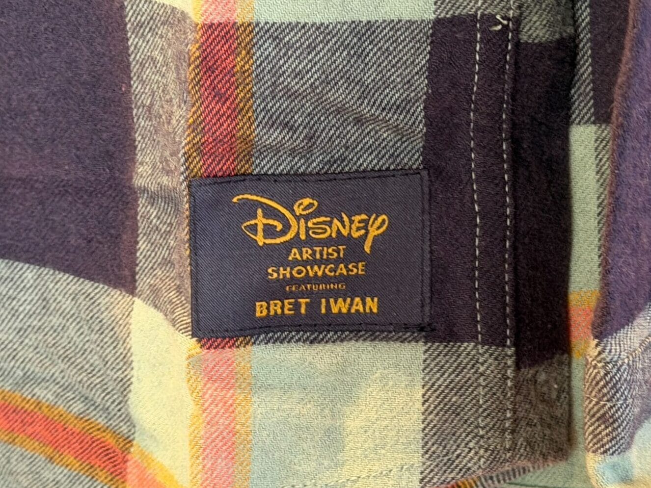 A detailed look at a plaid shirt from the Mickey Mouse Collection, showcasing Bret Iwan’s Disney Artist Showcase label—park exclusive.