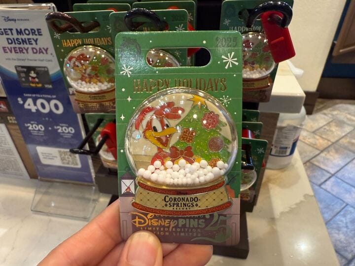 A hand displays a Walt Disney World Coronado Springs "Happy Holidays" pin featuring Daisy Duck inside a festive snow globe design.