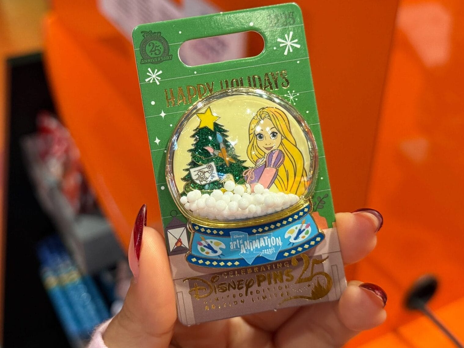 A hand displays a Walt Disney World holiday pin with Rapunzel, set against an orange store background—no castle imagery shown.