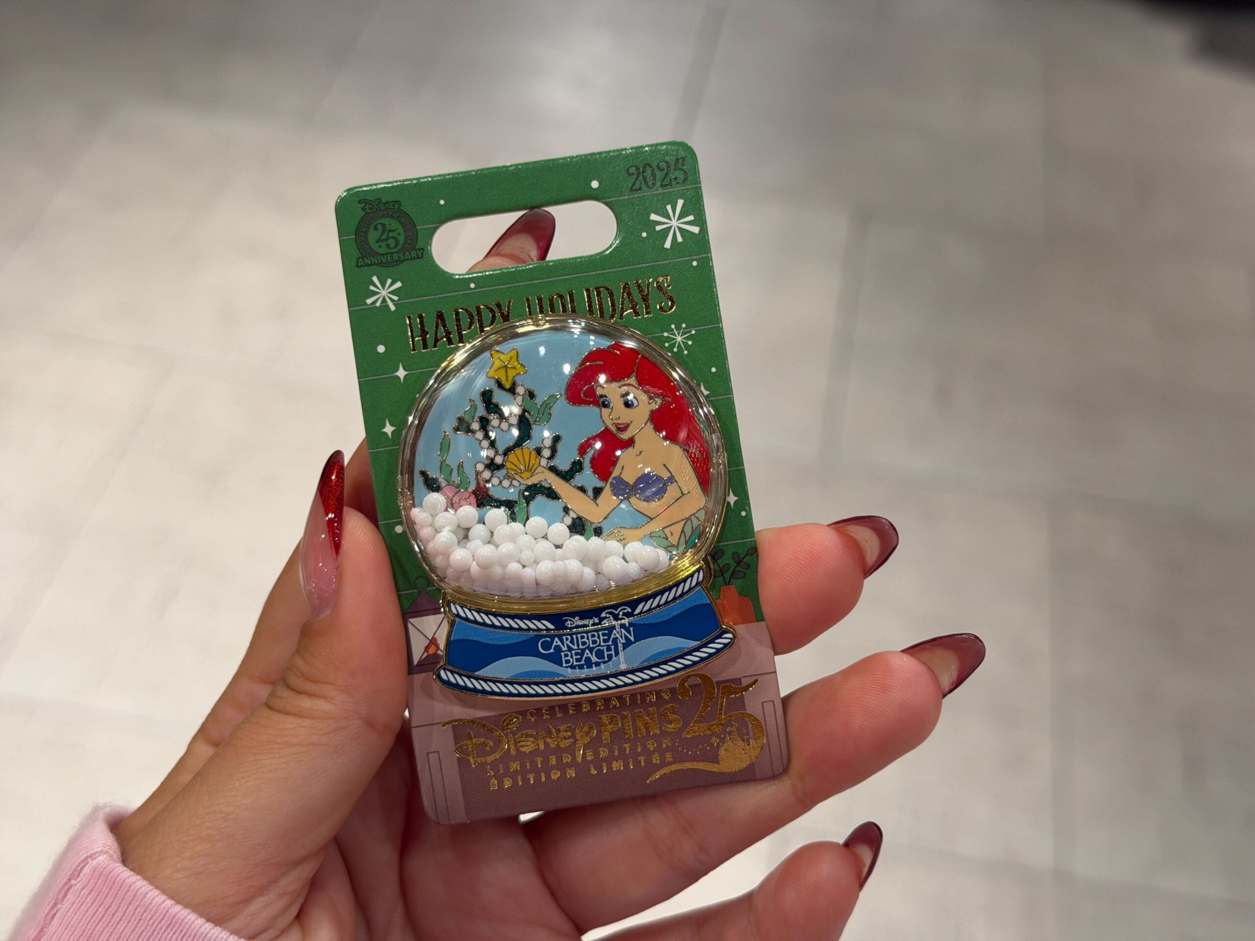 Hand holding a Walt Disney World holiday pin showing Ariel from The Little Mermaid in a snow globe with "Happy Holidays" text.