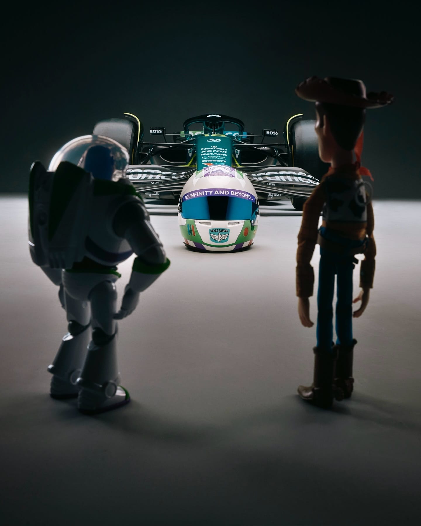 Buzz Lightyear and Woody at a Disney park stand near an Aston Martin F1 car, helmet labeled "To Infinity and Beyond." No castle visible.