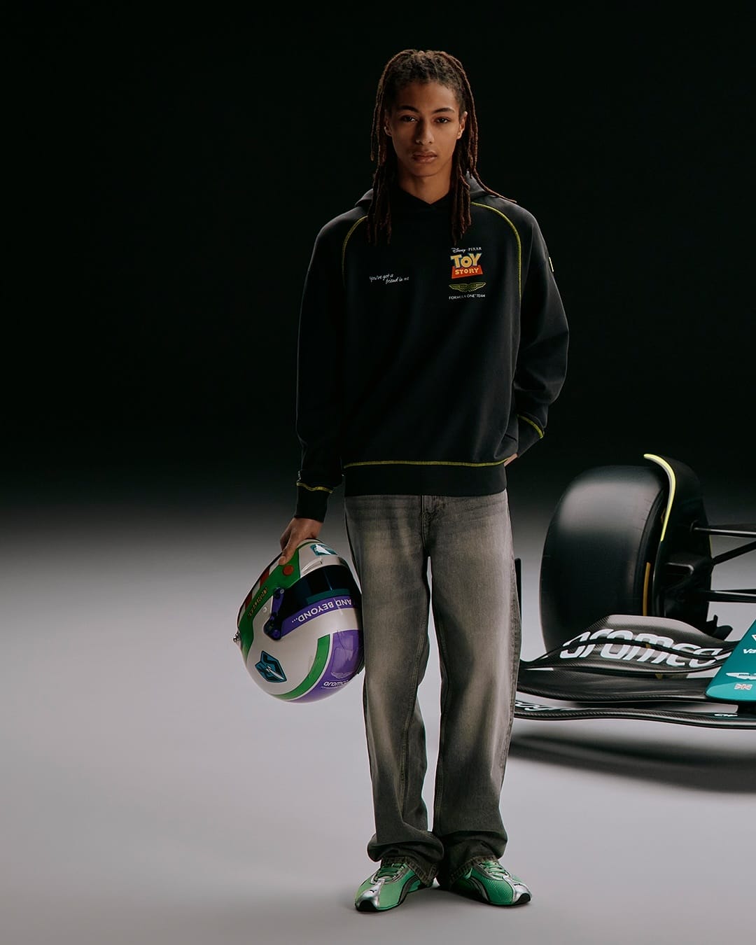 A person with long braids holds a racing helmet, standing by an Aston Martin car—reminiscent of Cars Land at Disneyland, on a dark background.