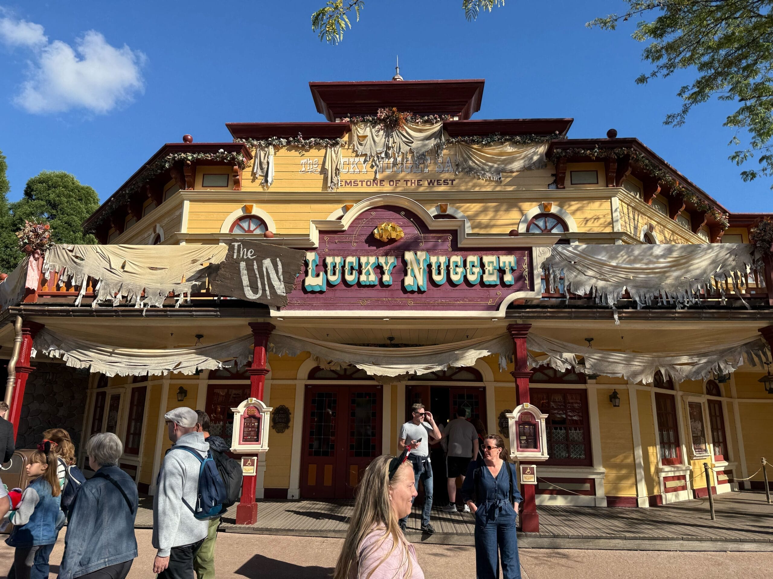 Guests stroll by the Lucky Nugget Saloon at Disneyland Paris, decked out for Halloween 2026 under bright blue skies. No castle in view.