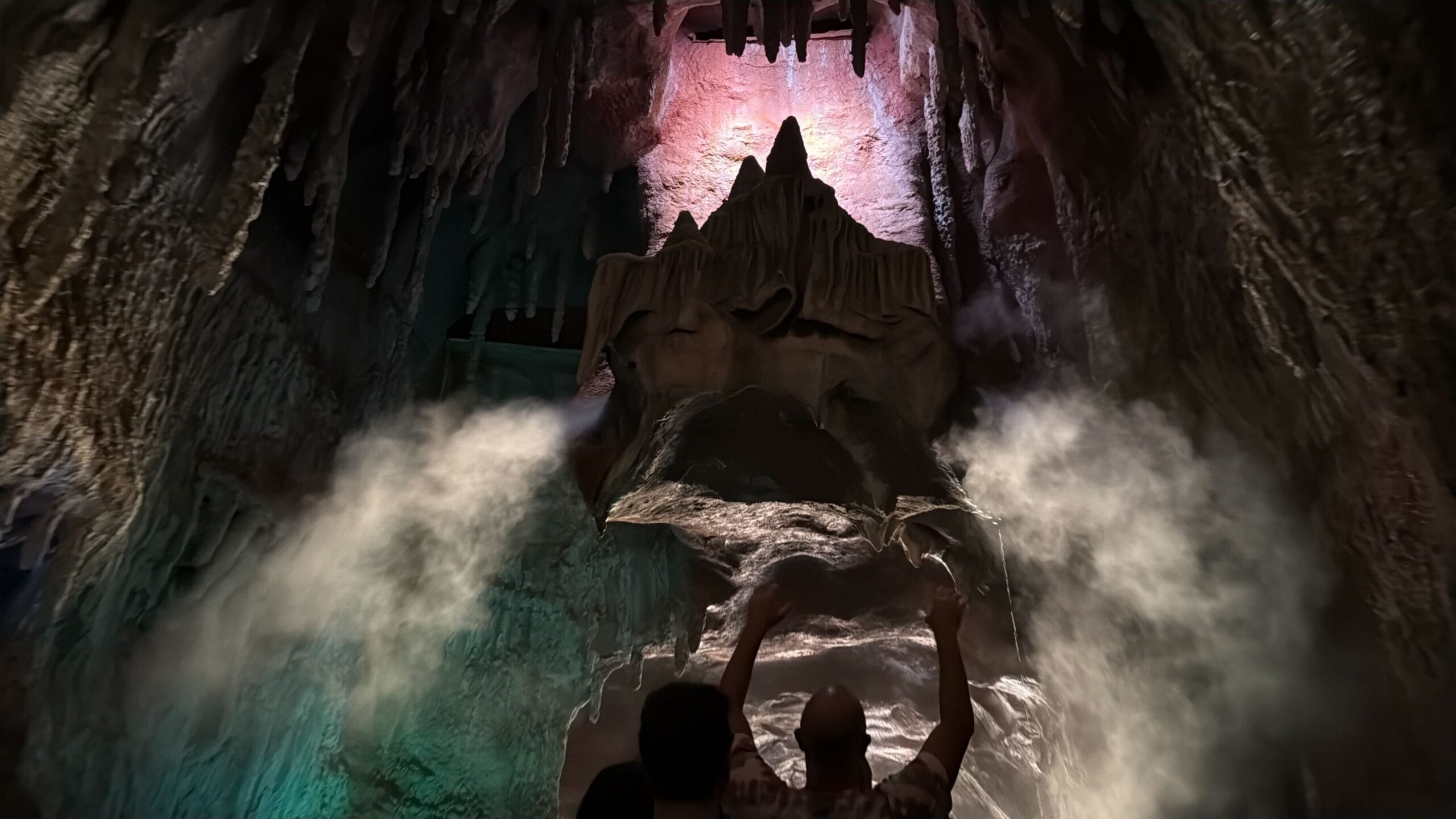 Two people raise their arms in a foggy cave with red and green lights, recalling Disneyland’s Big Thunder Mountain Railroad ambiance.