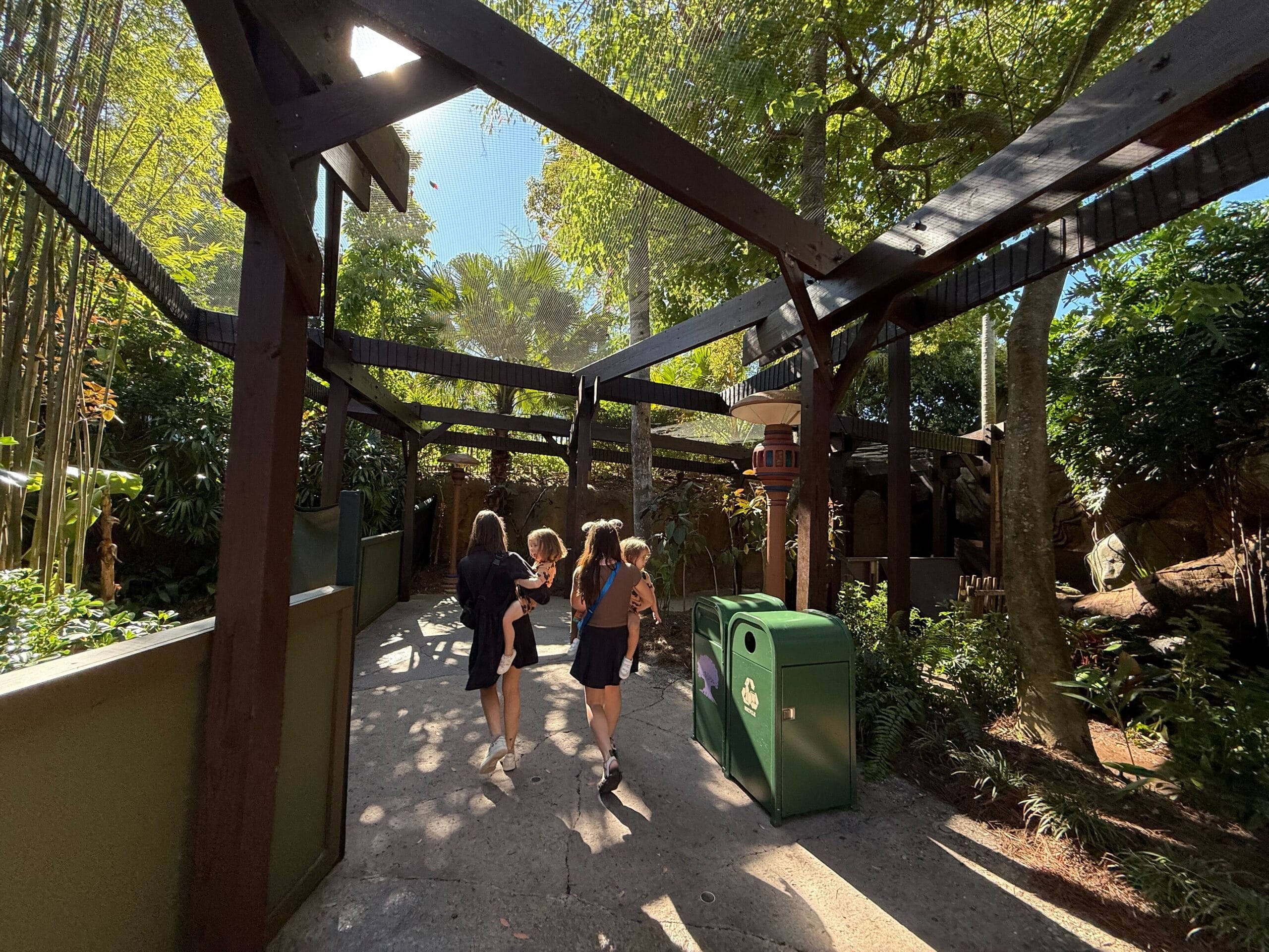 Three people stroll a shady, tree-lined walkway with wooden beams and a green Disney park trash can on a bright, sunny day.