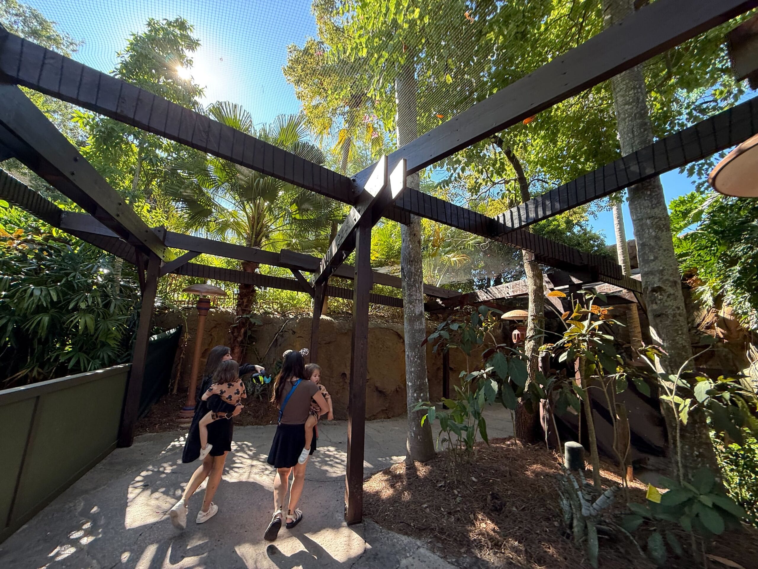 Three people stroll a sunlit pathway under wooden beams and lush greenery, evoking Disney park walkways near castle entrances.