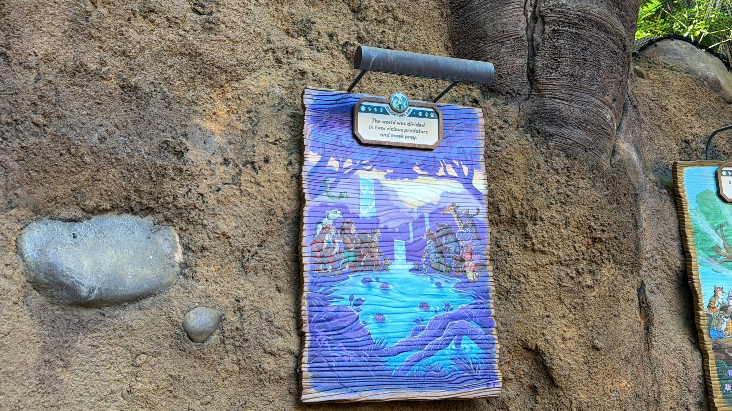 A vibrant sign on a rocky Animal Kingdom wall depicts animals by water under purple trees, evoking Zootopia vibes. No castle shown.