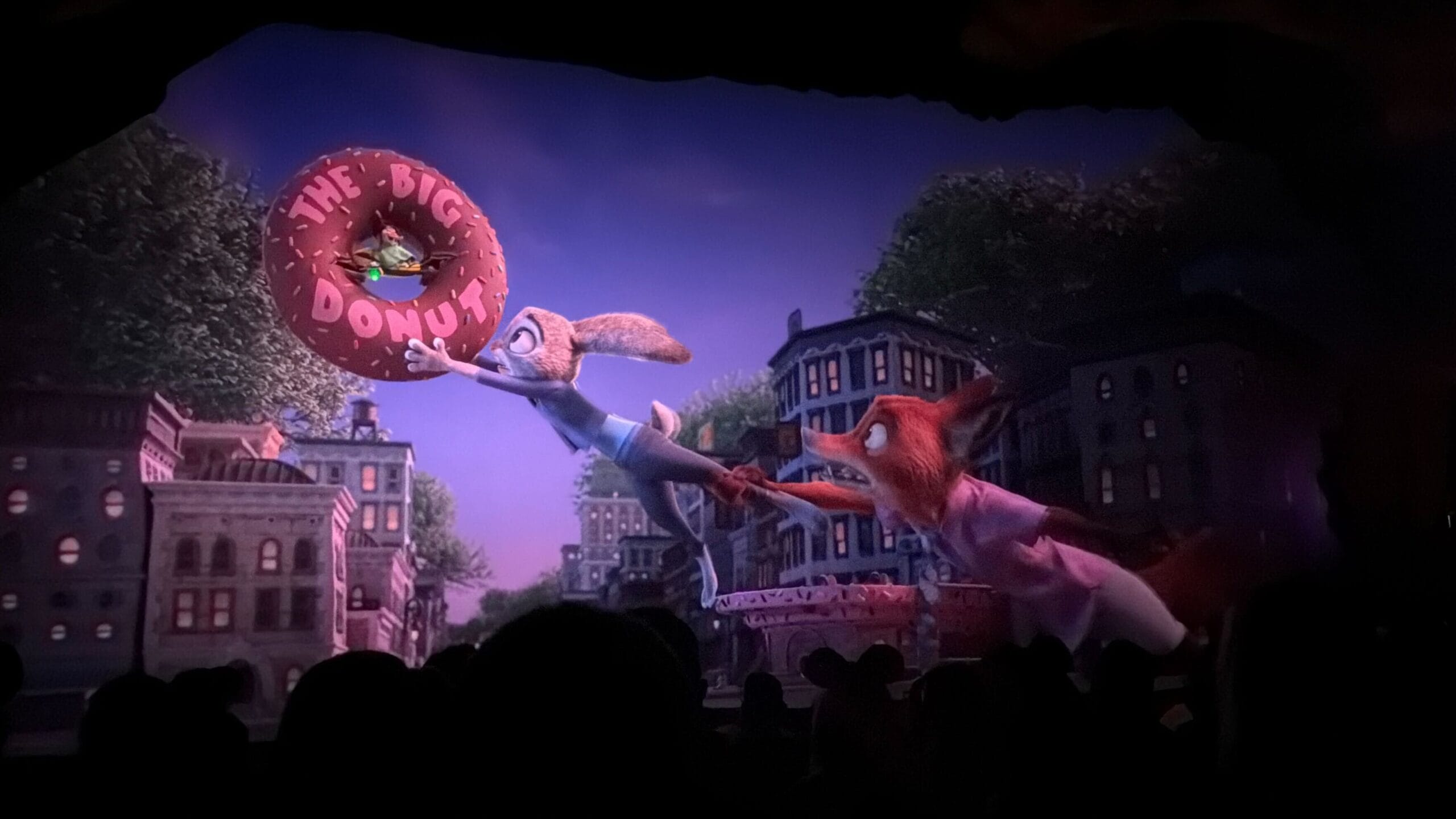 Animated Zootopia bunny and fox reach for a giant donut above city buildings, crowd below—no castle like those at Disney Parks.