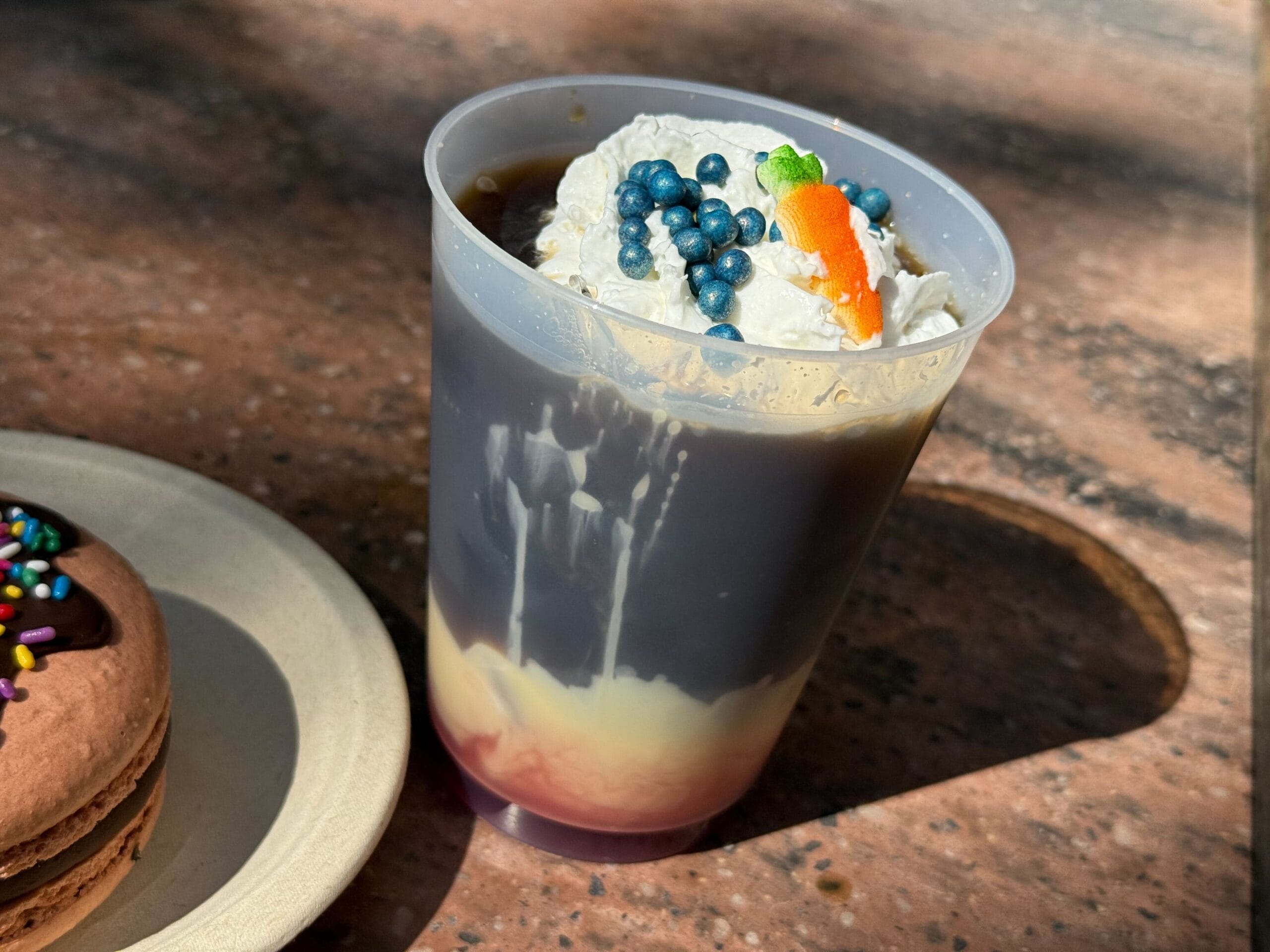 A magical iced coffee with cream, whipped cream, blue sprinkles, and a macaron beside it—just like a Disney Parks treat.
