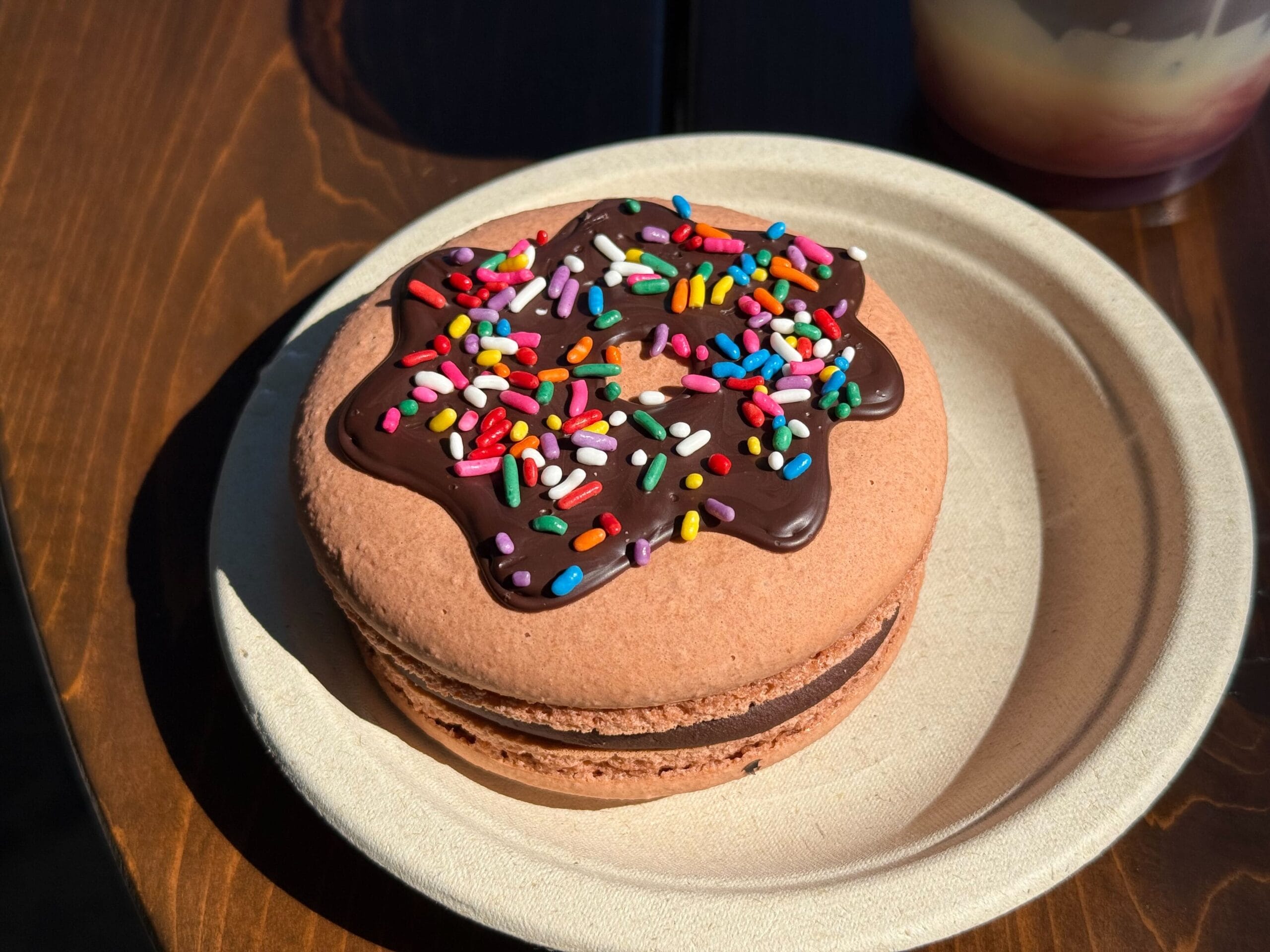 A chocolate macaron with chocolate icing and colorful sprinkles on a paper plate, perfect for a sweet treat at Disney parks.