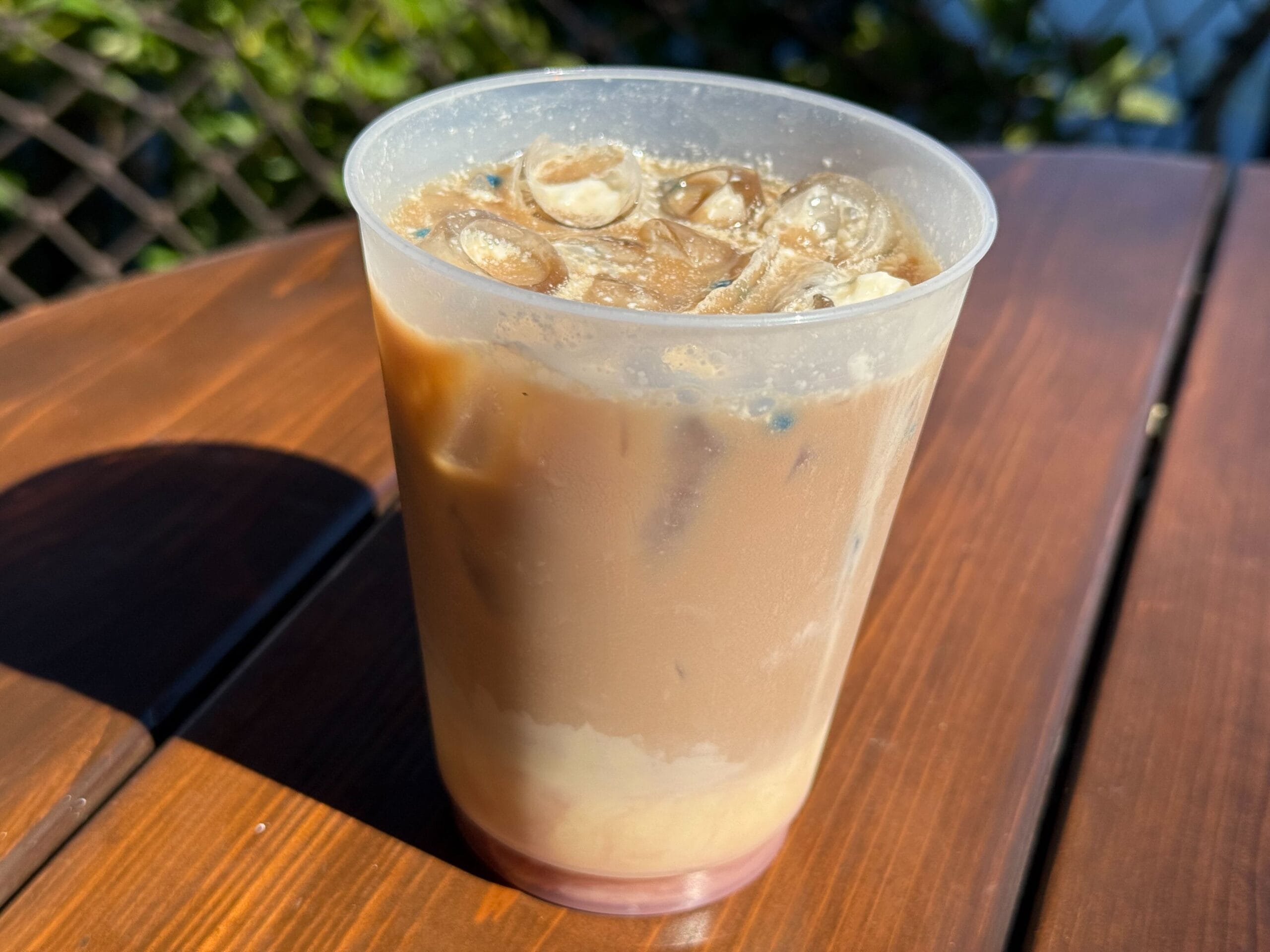 A plastic cup of iced coffee with milk rests on a wooden table in sunlight, reminiscent of Main Street cafés at Disneyland parks.