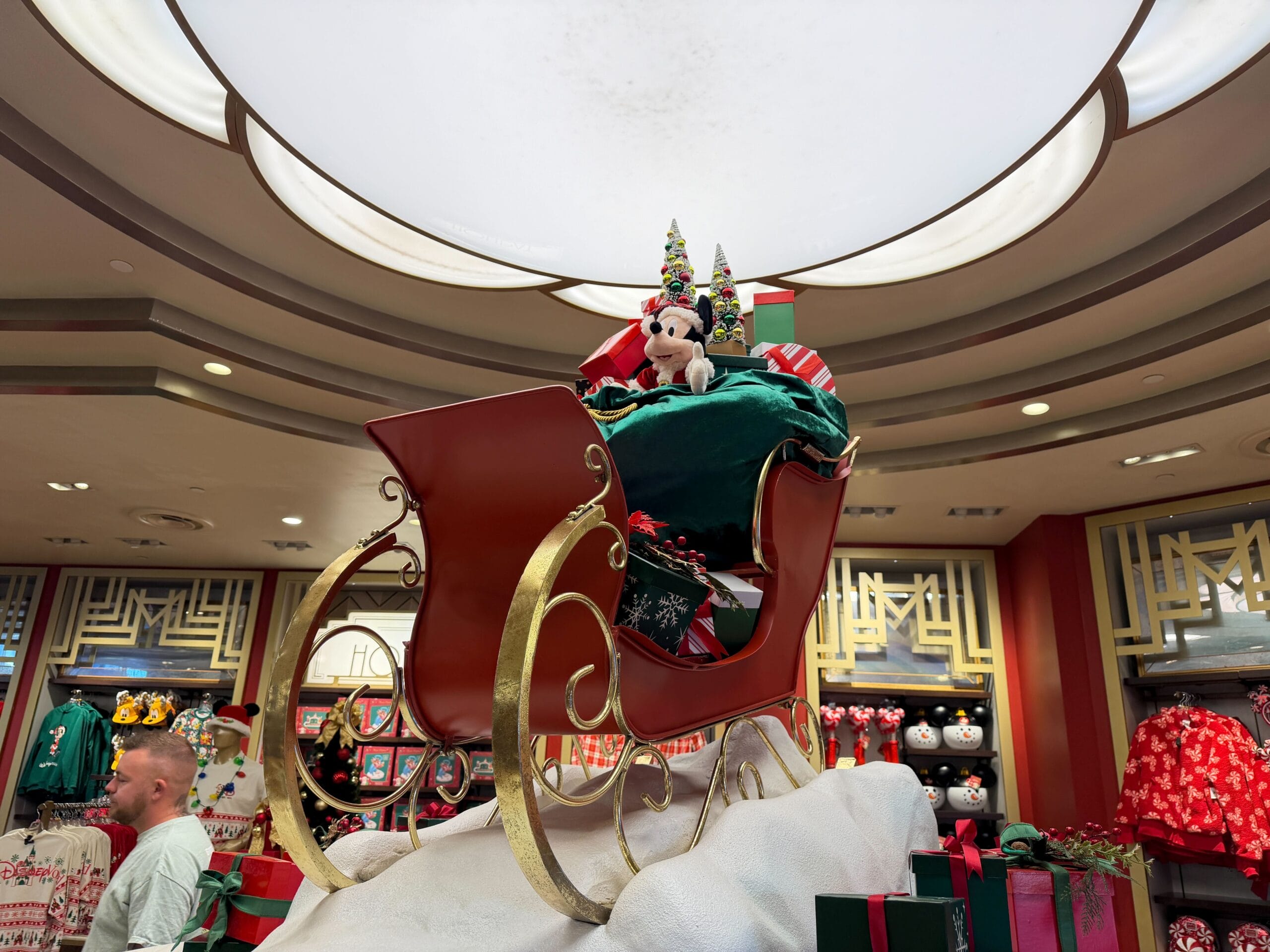 Red sleigh with gifts and a Mickey plush, surrounded by festive Disney park-style decor and circular lighting overhead indoors.