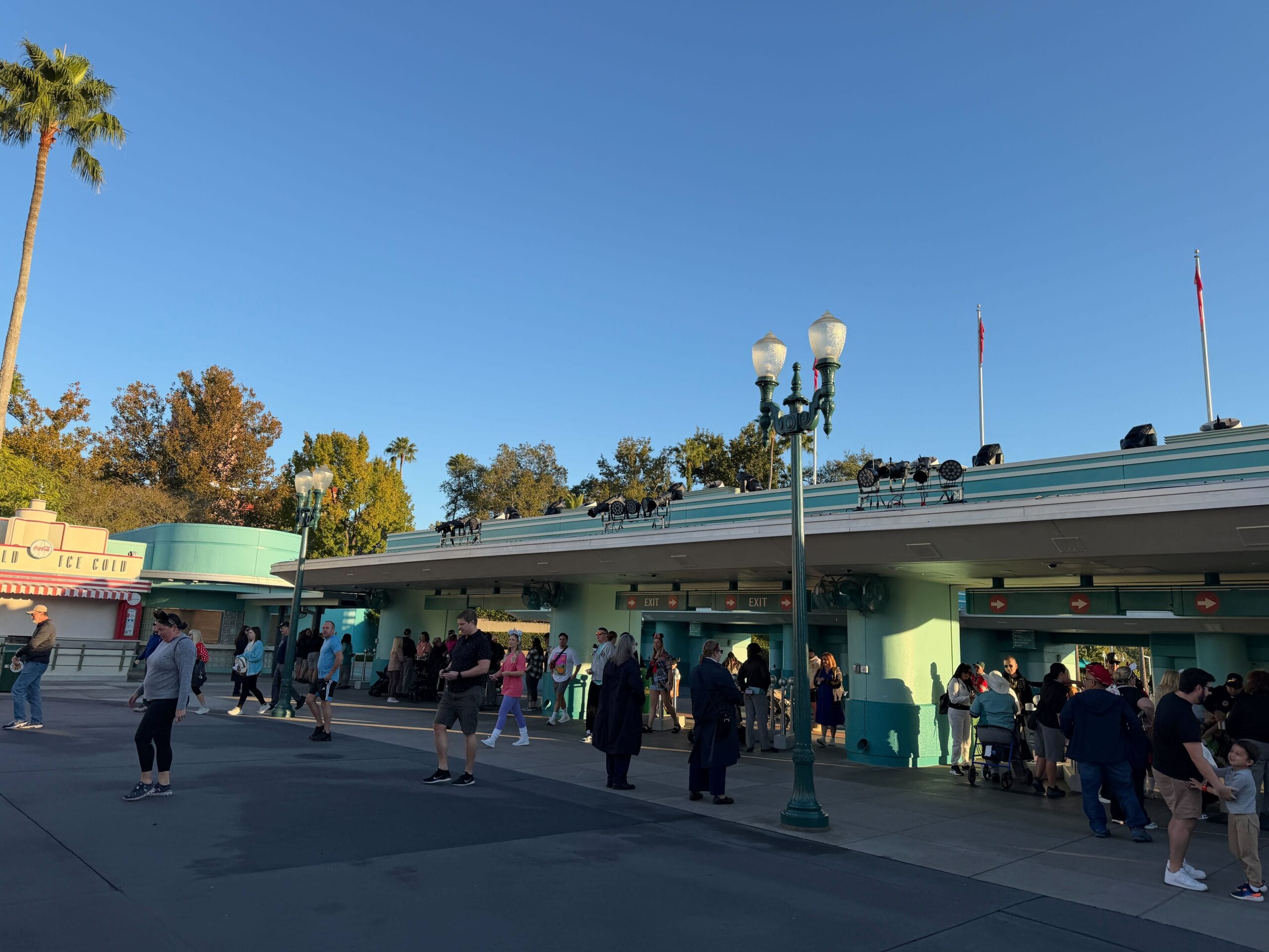 Guests stroll and gather by a teal ticket booth beneath a clear sky, near trees and lampposts reminiscent of Disney park entrances.