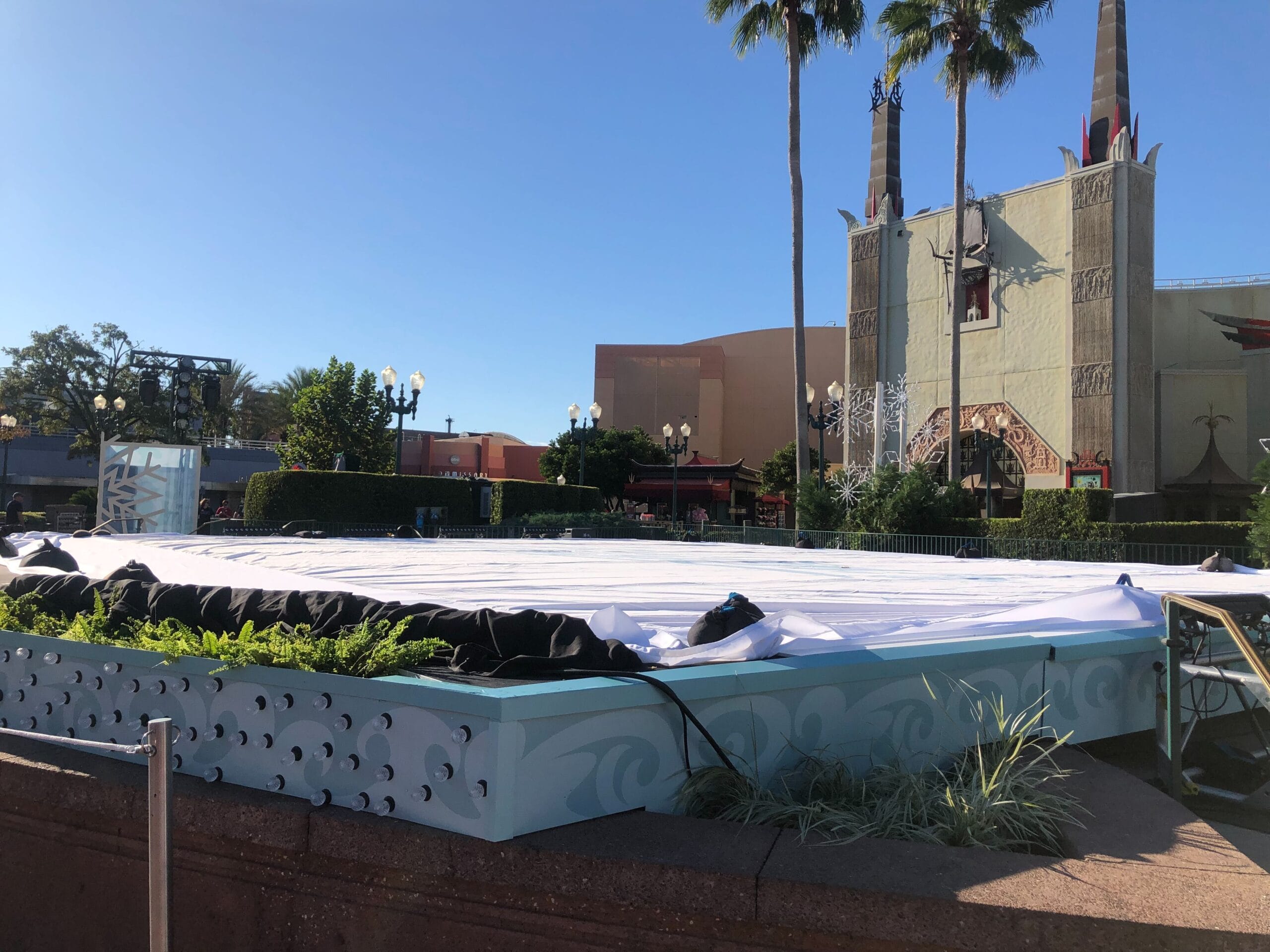 A large, covered stage outdoors near a grand, castle-like building reminiscent of Disney park facades, beneath a clear blue sky.