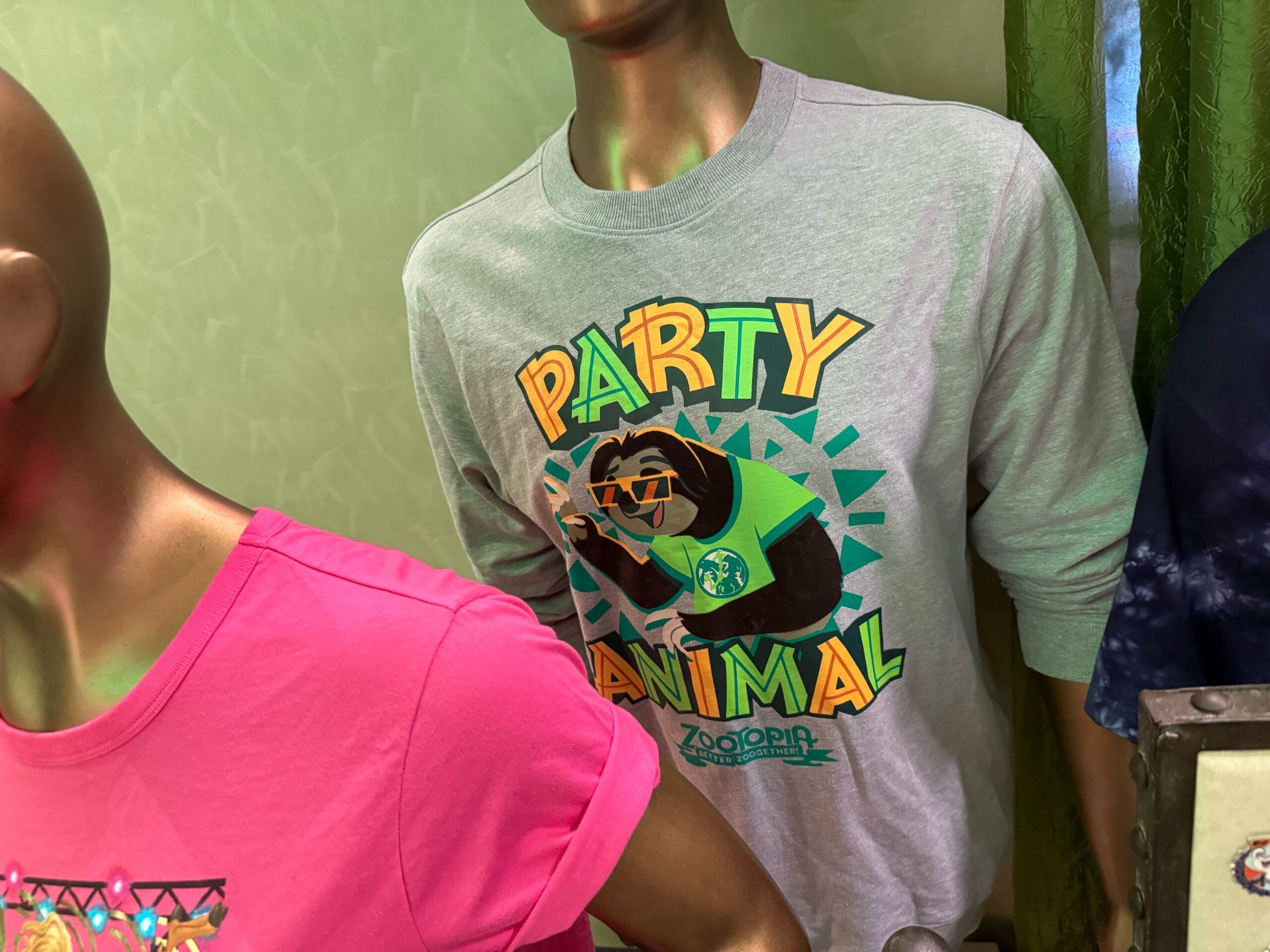 Two mannequins in a Disney Park shop: one wears a pink tee, the other a gray Zootopia sloth “Party Animal” sweatshirt.