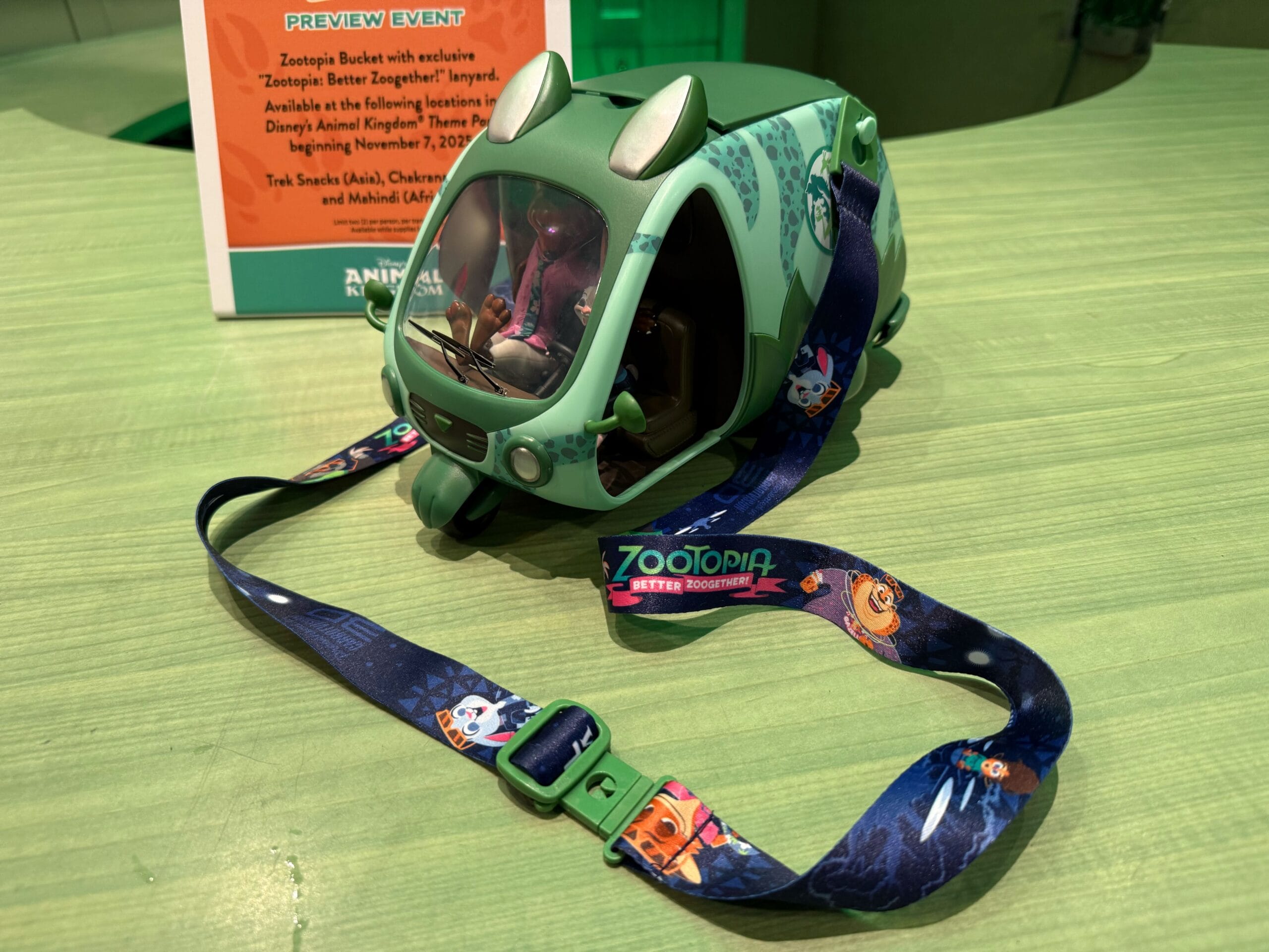 A green Zootopia popcorn bucket with a colorful lanyard sits on a light green table, fun merch seen at various Disney Parks.