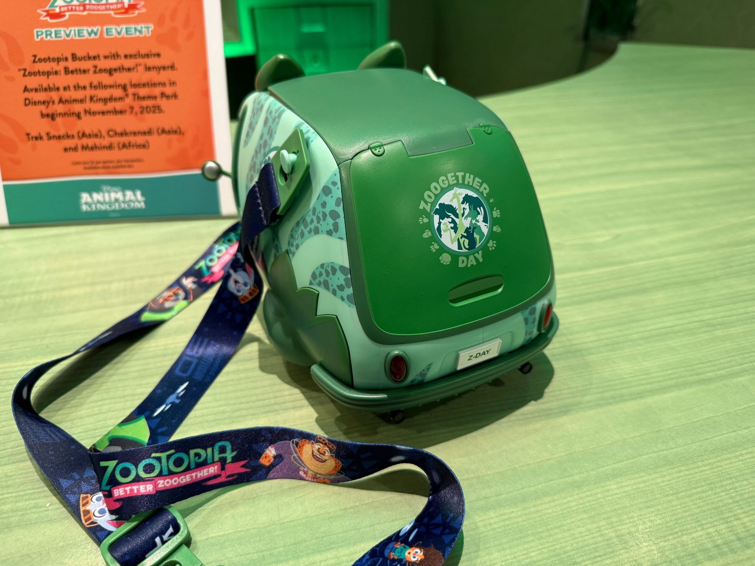 A green Zootopia popcorn bucket with a themed lanyard is displayed beside an orange Better Zoogether event sign at a Disney park.