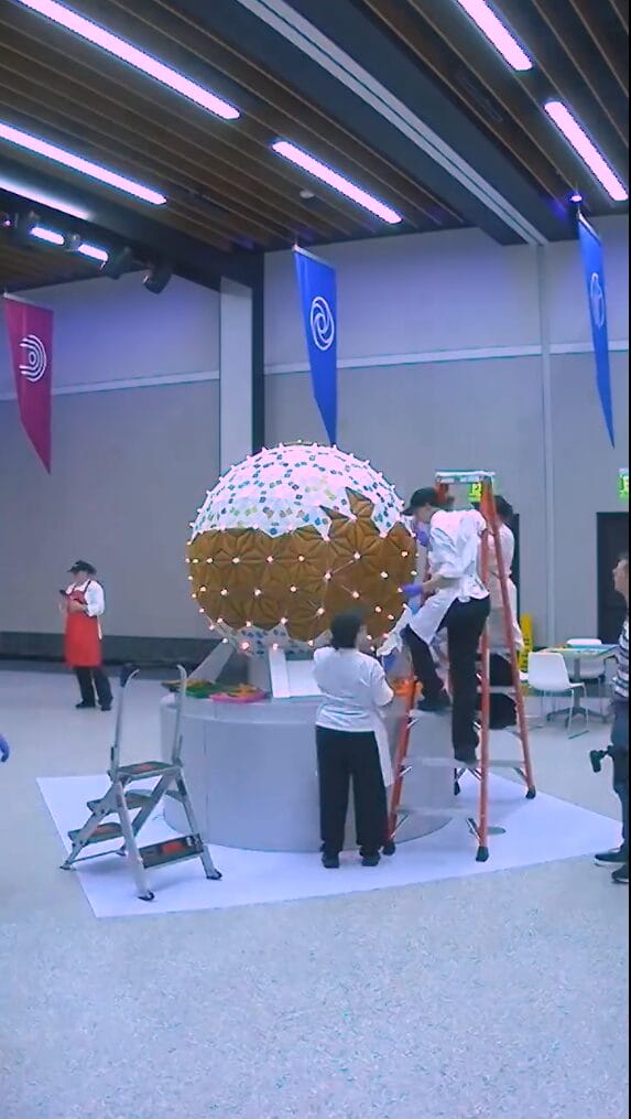 The video shows the team of pastry chefs assembling the display by attaching gingerbread panels to a lighted spherical base.