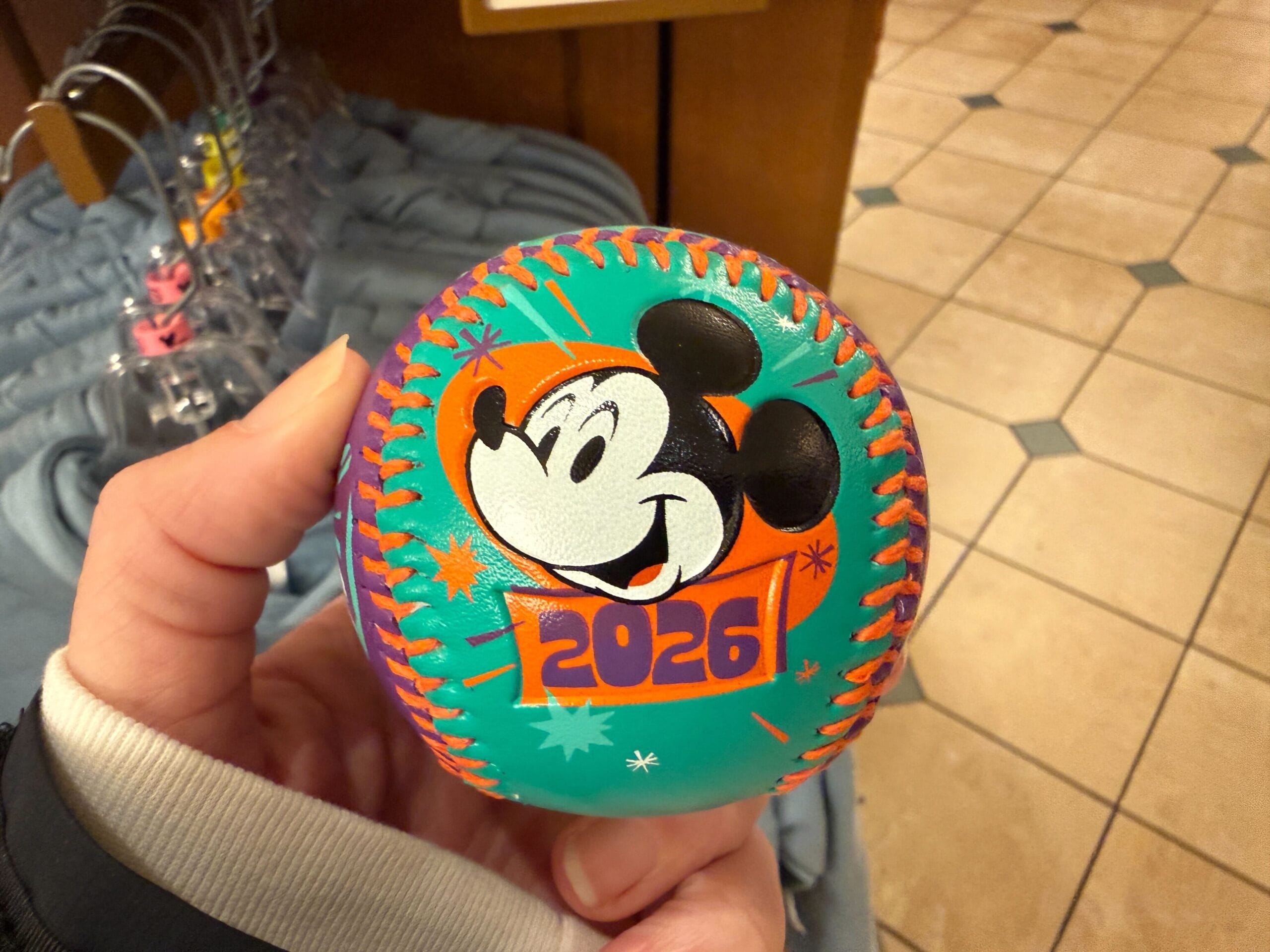 The baseball is aqua and purple with bright orange stitching. Mickey, Minnie, and theme park icons are pictured with stars.
