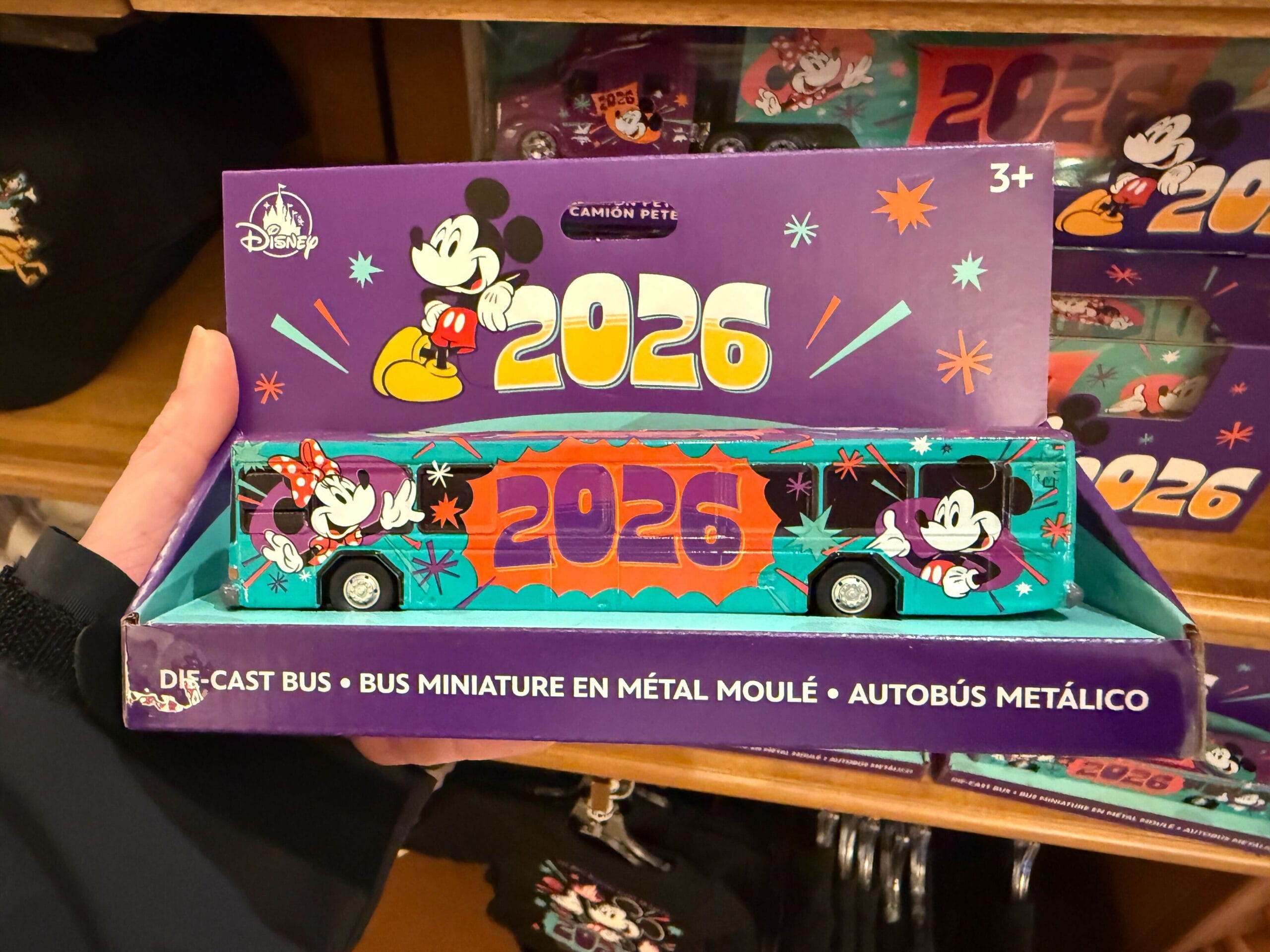 The bus is mainly aqua with a purple roof. "2026" is on the roof and both sides, between images of Mickey and Minnie.