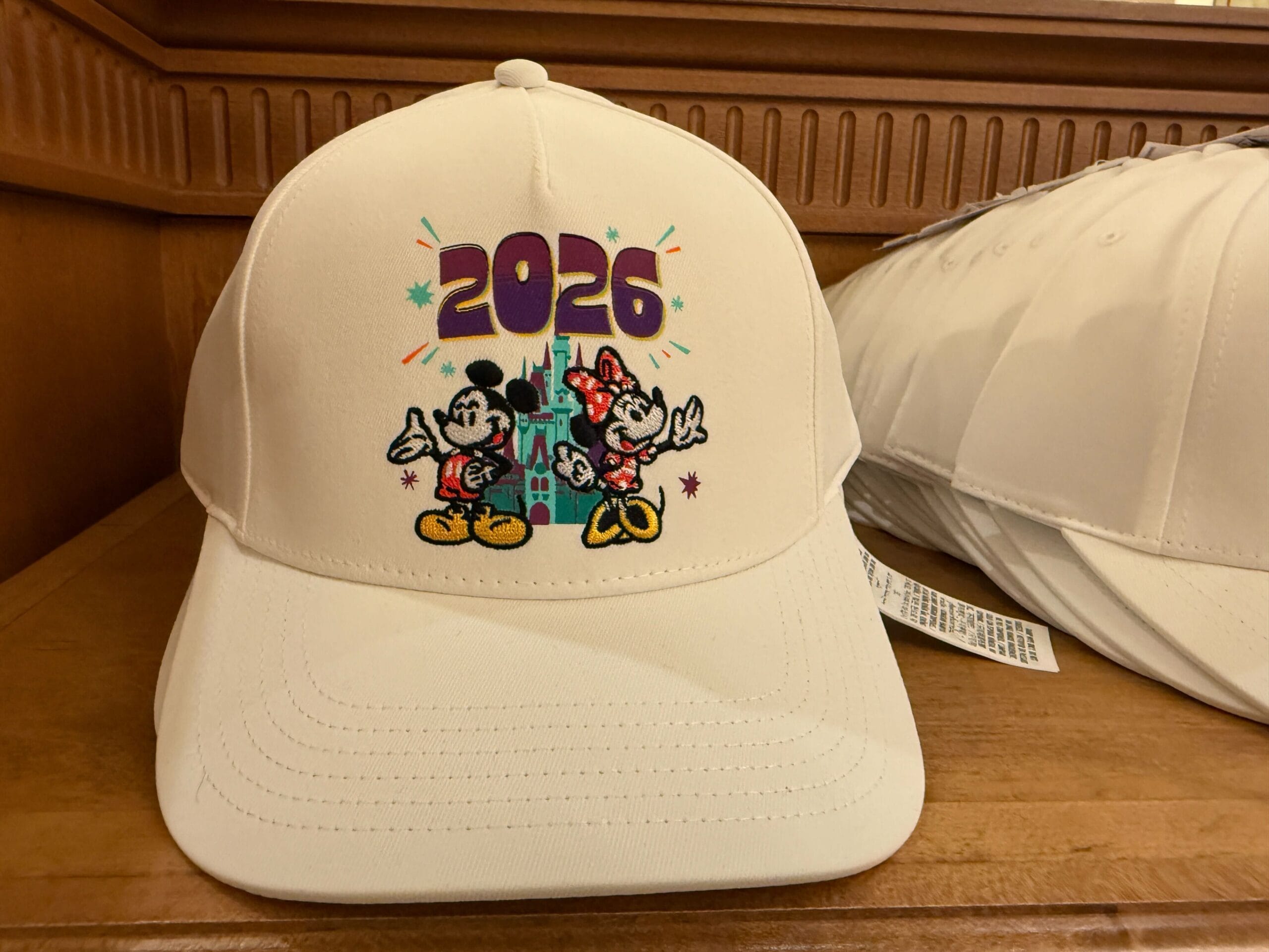 This white ball cap features an embroidered appliqué of Mickey, Minnie, Cinderella Castle, and the year in purple.