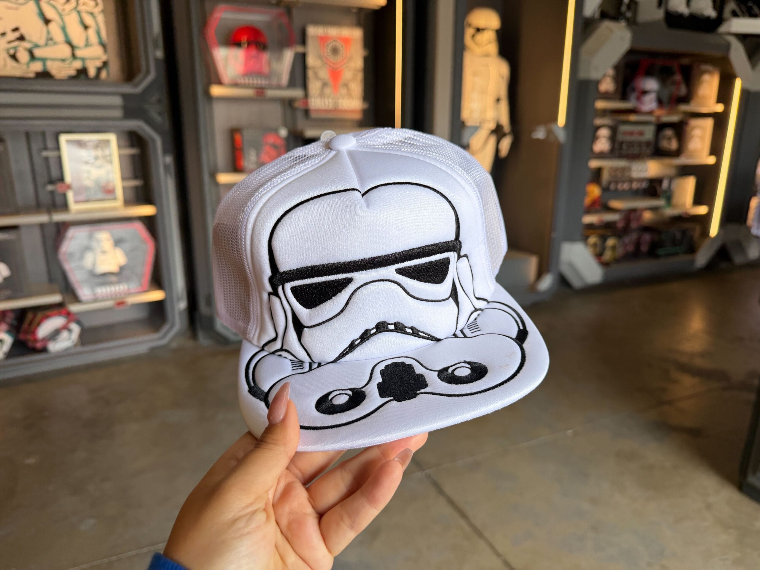 This white cap has a fabric front panel and fabric-covered brim, and a plastic mesh back half. Embroidered in black across the front and brim are the details of a Stormtrooper helmet.